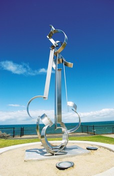 Photograph of a large stainless steel sculpture resembling clouds, wind and waves and set onto a concrete base. The base is set within a round sand area, the bay, sky and clouds are in the background.