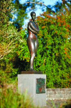 Photograph of an outdoor sculpture of a female, cast in bronze. Figure has crossed arms and is wearing bathers and a swimming cap. Stands on a polished granite pedestal, to which is affixed a low relief bronze sculptural panel.