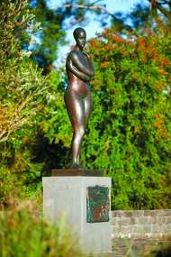 Photograph of an outdoor sculpture of a female, cast in bronze. Figure has crossed arms and is wearing bathers and a swimming cap. Stands on a polished granite pedestal, to which is affixed a low relief bronze sculptural panel.