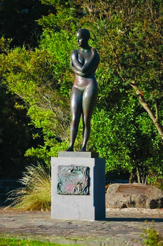 Photograph of an outdoor sculpture of a female, cast in bronze. Figure has crossed arms and is wearing bathers and a swimming cap. Stands on a polished granite pedestal, to which is affixed a low relief bronze sculptural panel.