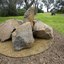 Photograph of an outdoor sculpture comprising of a group of six large bluestones laid on crushed yellow ochre sandstone surrounded by grass and distant vegetation.