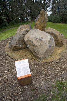 Photograph of an outdoor sculpture comprising of a group of six large bluestones laid on crushed yellow ochre sandstone surrounded by grass and distant vegetation.