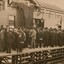 Sepia photograph of a crowd of largely men in suits and hats and some women with long dresses and hats. They are standing on a wooden train platform in front of a station building, looking away from the camera towards a man without hat appearing to deliver a speech. There is a large flag or banner hanging from the centre of the building and bunting on either side. 