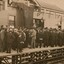 Sepia photograph of a crowd of largely men in suits and hats and some women with long dresses and hats. They are standing on a wooden train platform in front of a station building, looking away from the camera towards a man without hat appearing to deliver a speech. There is a large flag or banner hanging from the centre of the building and bunting on either side. 