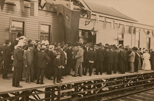 Sepia photograph of a crowd of largely men in suits and hats and some women with long dresses and hats. They are standing on a wooden train platform in front of a station building, looking away from the camera towards a man without hat appearing to deliver a speech. There is a large flag or banner hanging from the centre of the building and bunting on either side. 