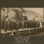 Sepia photograph of a crowd of largely men in suits and hats and some women with long dresses and hats. They are standing on a wooden train platform in front of a station building, looking away from the camera towards a man without hat appearing to deliver a speech. There is a large flag or banner hanging from the centre of the building and bunting on either side.  Photograph has a grey mount with white lettering below it.