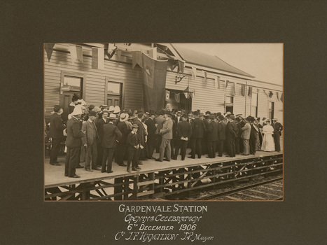 Sepia photograph of a crowd of largely men in suits and hats and some women with long dresses and hats. They are standing on a wooden train platform in front of a station building, looking away from the camera towards a man without hat appearing to deliver a speech. There is a large flag or banner hanging from the centre of the building and bunting on either side.  Photograph has a grey mount with white lettering below it.