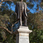 Bronze statue of a larger than life-sized body of a suited gentleman standing on a tapered granite pedestal. One hand holds a scroll, the other hand is outstretched. Sculpture is outdoors with gum trees visible in background.