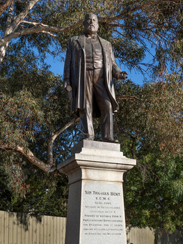 Bronze statue of a larger than life-sized body of a suited gentleman standing on a tapered granite pedestal. One hand holds a scroll, the other hand is outstretched. Sculpture is outdoors with gum trees visible in background.