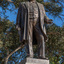 Bronze statue of a larger than life-sized body of a suited gentleman standing on a tapered granite pedestal. One hand holds a scroll, the other hand is outstretched. Sculpture is outdoors with gum trees visible in background.