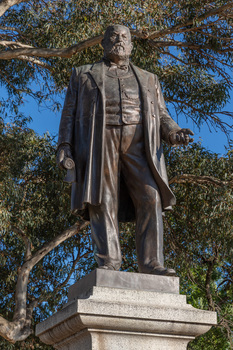 Bronze statue of a larger than life-sized body of a suited gentleman standing on a tapered granite pedestal. One hand holds a scroll, the other hand is outstretched. Sculpture is outdoors with gum trees visible in background.