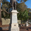 Bronze statue of a larger than life-sized body of a suited gentleman standing on a tapered granite pedestal. One hand holds a scroll, the other hand is outstretched. Sculpture is outdoors and surrounded by brick pavers, gum trees and fence visible in background. A drinking fountain sculpture can be seen on the far right.