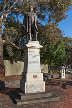 Bronze statue of a larger than life-sized body of a suited gentleman standing on a tapered granite pedestal. One hand holds a scroll, the other hand is outstretched. Sculpture is outdoors and surrounded by brick pavers, gum trees and fence visible in background. A drinking fountain sculpture can be seen on the far right.