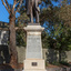 Bronze statue of a larger than life-sized body of a suited gentleman standing on a tapered granite pedestal. One hand holds a scroll, the other hand is outstretched. Sculpture is outdoors and surrounded by brick pavers, gum trees and fence visible in background. 