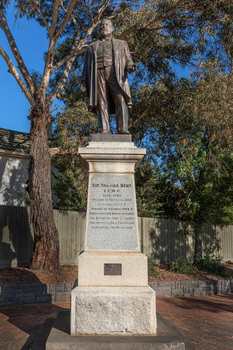 Bronze statue of a larger than life-sized body of a suited gentleman standing on a tapered granite pedestal. One hand holds a scroll, the other hand is outstretched. Sculpture is outdoors and surrounded by brick pavers, gum trees and fence visible in background. 