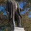 Bronze statue of a larger than life-sized body of a suited gentleman standing on a tapered granite pedestal. Sculpture is outdoors with gum trees visible in background.