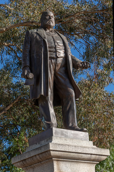 Bronze statue of a larger than life-sized body of a suited gentleman standing on a tapered granite pedestal. Sculpture is outdoors with gum trees visible in background.