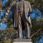 Bronze statue of a larger than life-sized body of a suited gentleman standing on a tapered granite pedestal. Sculpture is outdoors with gum trees visible in background.