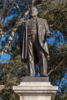Bronze statue of a larger than life-sized body of a suited gentleman standing on a tapered granite pedestal. Sculpture is outdoors with gum trees visible in background.