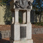 A granite drinking fountain which has a square base, with 4 small pillars supporting a curved canopy which contains an oval bronze relief medallion of a woman, from shoulders up, flanked by 2 cherubs.  The outdoor drinking fountain sits on a bluestone base surrounded by brick pavers, a garden bed, leaves and a fence are visible in the background.