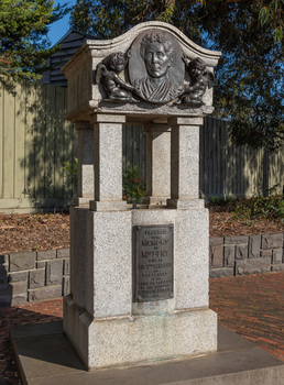 A granite drinking fountain which has a square base, with 4 small pillars supporting a curved canopy which contains an oval bronze relief medallion of a woman, from shoulders up, flanked by 2 cherubs.  The outdoor drinking fountain sits on a bluestone base surrounded by brick pavers, a garden bed, leaves and a fence are visible in the background.