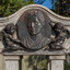 Detail of an oval bronze relief medallion of a woman, from shoulders up flanked by 2 cherubs.  The medallion is attached to a granite canopy which sits on four small plinths. 