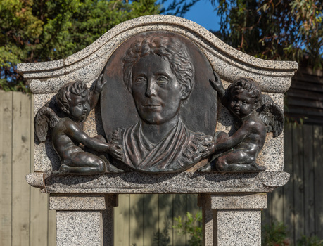 Detail of an oval bronze relief medallion of a woman, from shoulders up flanked by 2 cherubs.  The medallion is attached to a granite canopy which sits on four small plinths. 