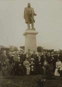 A group of seated and standing men and women in front of the large bronze statue of Sir Thomas Bent, which sits on a tall granite pedestal.