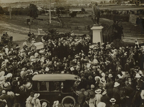 A large group of people surrounding a small memorial fountain on the left which is being unveiled by the Mayoress. To the right is the large bronze statue of Sir Thomas Bent which stands on a tall granite pedestal. In the foreground is an early 20th century car. Paddocks and houses can be seen in the distance.