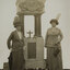 Two women in hats, long skirts and jackets, stand either side of a granite memorial fountain which is a square base on top sits four rounded pillars which support a canopy. A bronzed relief medallion of a portrait of a woman, flanked by two cherubs is on the canopy.