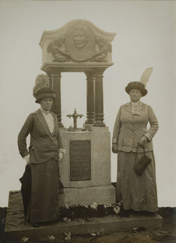 Two women in hats, long skirts and jackets, stand either side of a granite memorial fountain which is a square base on top sits four rounded pillars which support a canopy. A bronzed relief medallion of a portrait of a woman, flanked by two cherubs is on the canopy.