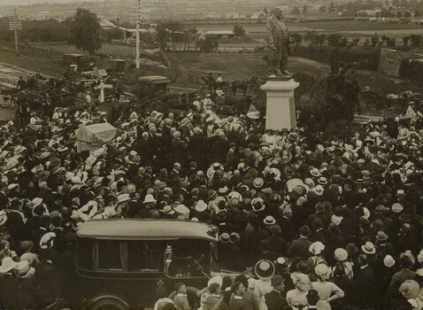 A large group of people surrounding the large bronze statue of Sir Thomas Bent which stands on a tall granite pedestal. On the left of this is another smaller memorial which is covered by a cloth. In the foreground is an early 20th century car. Paddocks and houses can be seen in the distance.