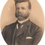 Oval shaped sepia toned portrait photograph of a man from chest up in a suit with dark hair in a deep side part, facial hair, a handkerchief in right hand pocket of suit jacket and a dark tie.