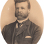 Oval shaped sepia toned portrait photograph of a man from chest up in a suit with dark hair in a deep side part, facial hair, a handkerchief in right hand pocket of suit jacket and a dark tie.