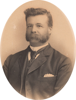 Oval shaped sepia toned portrait photograph of a man from chest up in a suit with dark hair in a deep side part, facial hair, a handkerchief in right hand pocket of suit jacket and a dark tie.