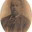 Oval shaped sepia toned portrait photograph of a man from chest up with a receding hairline, a goatee, and wearing a suit with a tie