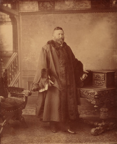 Photographic portrait of a standing bearded man of large stature, wearing mayoral robes with fur trims stands next to an ornately carved chair and desk. In the background is a set of stairs