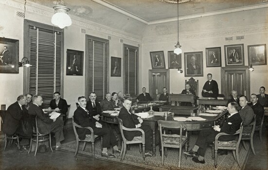 Gelatin silver photograph of the last council meeting held in the Brighton Town Hall. Councillors are seated around a large U-shaped council table surrounded by portraits of former Mayors.