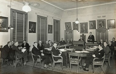Gelatin silver photograph of the last council meeting held in the Brighton Town Hall. Councillors are seated around a large U-shaped council table surrounded by portraits of former Mayors.
