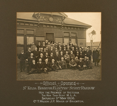 Sepia toned photograph of a group of men, standing and sitting, posing in front of a building with lawn in front. Mostly have their gaze towards the left of the camera.