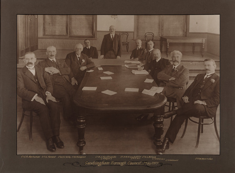 Sepia photograph of the a meeting of 10 suited men around a table. The man in the centre is standing