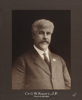 Sepia photograph of a formal portrait of seated man, chest up wearing grey suit, white collared shirt and dark tie and with Australian coat of arms pin on lapel. 