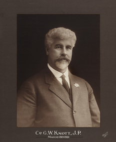 Sepia photograph of a formal portrait of seated man, chest up wearing grey suit, white collared shirt and dark tie and with Australian coat of arms pin on lapel. 