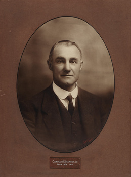 Oval shaped sepia photograph of a formal portrait of a seated man, chest up, wearing 3 piece suit, white collared shirt and tie.