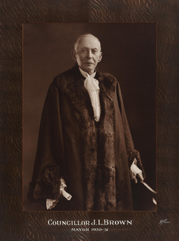 Sepia photograph of a formal portrait of standing man with short white hair and moustache wearing mayoral robes with fur trims and lace cuffs and jabot. 