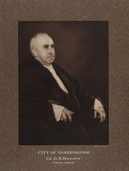 Sepia photograph of formal portrait of seated man with short grey hair, turned sideways slightly leaning back wearing mayoral robes with fur trims and lace cuffs and jabot