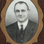 Black and white photograph of an oval portrait of a suited man with dark hair, surrounded by gold border
