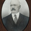 Black and white photograph of an oval portrait of a suited man with balding white hair and dark moustache