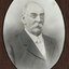 Black and white photograph of an oval portrait of a suited man with balding white hair, moustache and small spectacles.