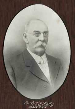 Black and white photograph of an oval portrait of a suited man with balding white hair, moustache and small spectacles.